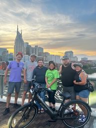 Six cyclists smiling by their bikes on a bridge at sunset with the Nashville skyline and twin‑spired skyscraper in the background