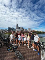 Group of people smiling with rental bikes on a bridge over the Cumberland River, downtown Nashville skyline with a twin‑spired high-rise under a dramatic cloud-filled blue sky.