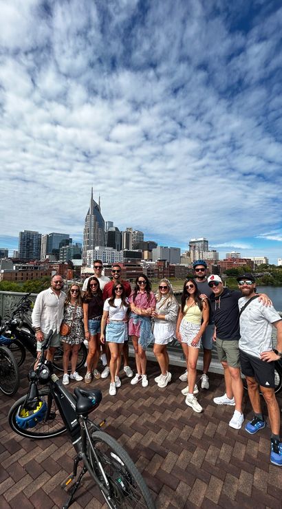 Group of people smiling with rental bikes on a bridge over the Cumberland River, downtown Nashville skyline with a twin‑spired high-rise under a dramatic cloud-filled blue sky.