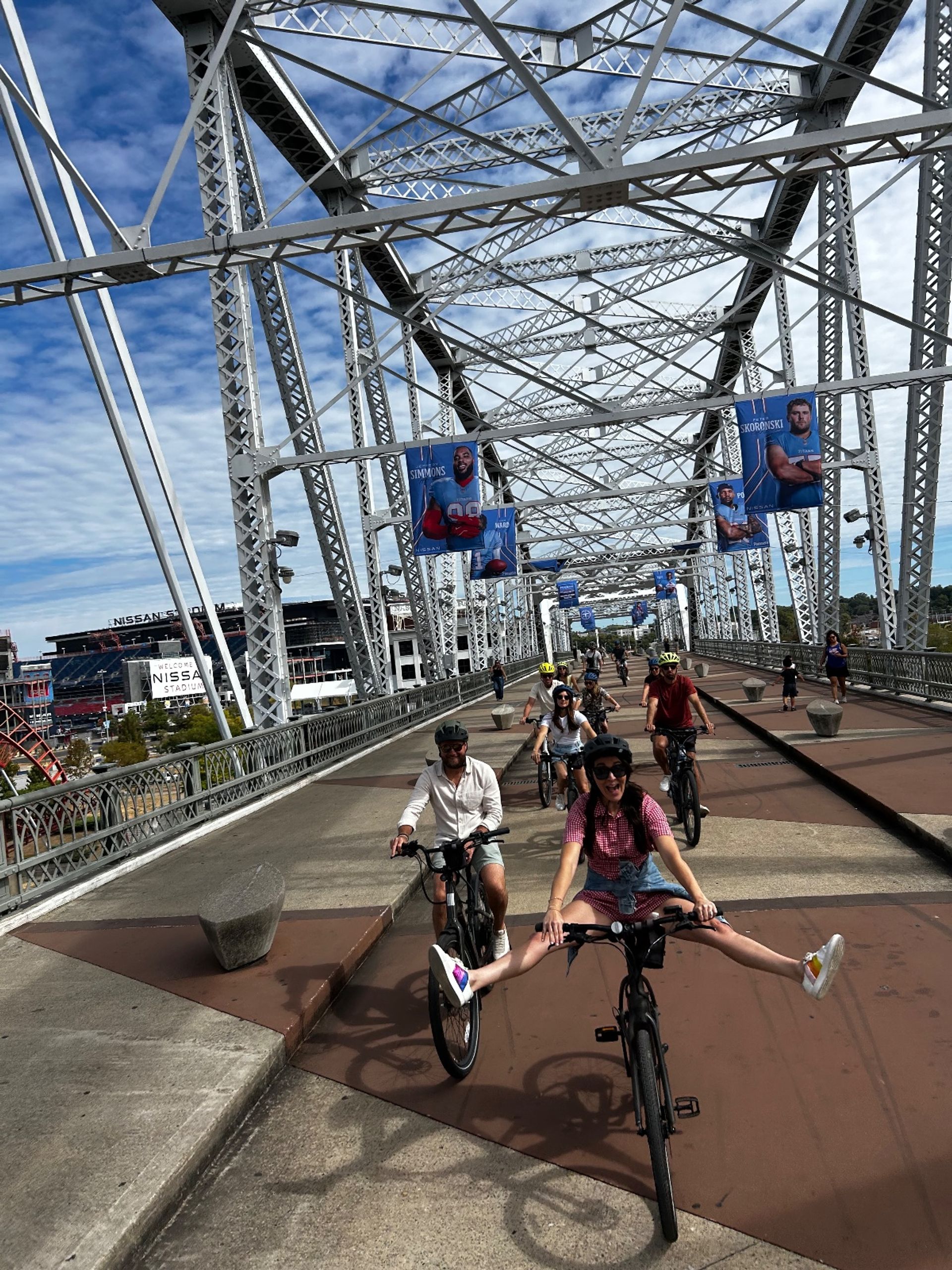 Group of cyclists riding across a large white steel-truss pedestrian bridge over a river, athlete banners hanging from the structure and a city stadium visible in the background under a bright blue sky; one rider playfully pedals with legs outstretched.