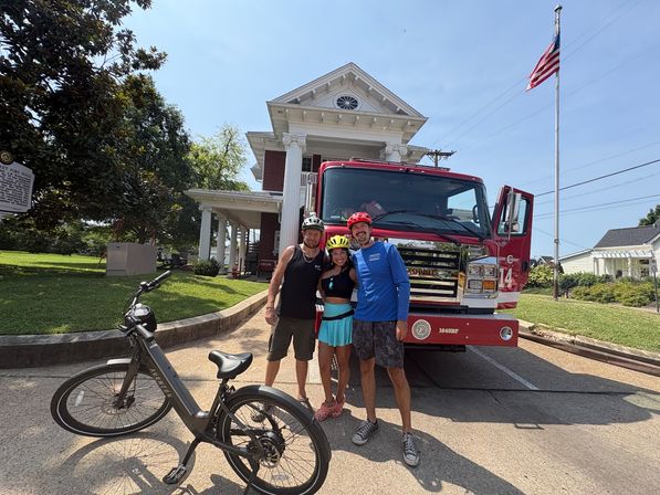 Three helmeted cyclists smiling in front of a red fire truck and white-columned historic firehouse, e-bike in foreground, American flag overhead on a sunny day