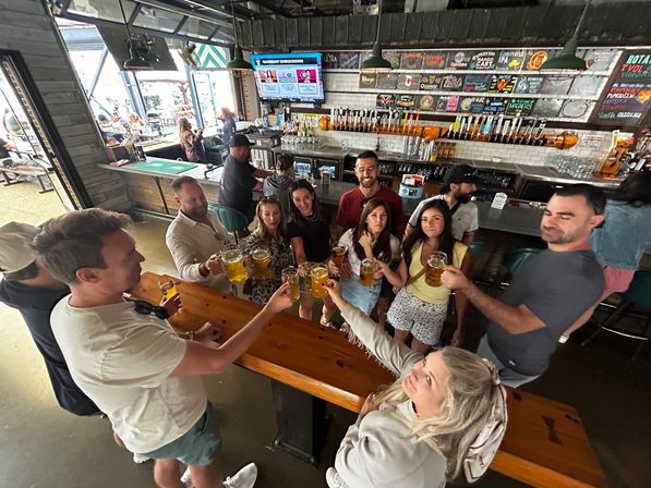 Group of friends clinking draft beer mugs at a lively taproom bar by a long wooden communal table, wall of taps and chalkboard beer menu behind, with open-air windows and outdoor seating visible