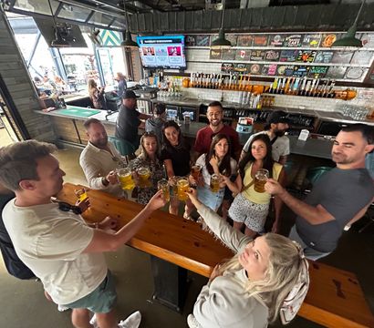 Group of friends clinking draft beer mugs at a lively taproom bar by a long wooden communal table, wall of taps and chalkboard beer menu behind, with open-air windows and outdoor seating visible