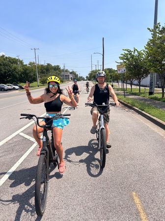 Group bike ride on an urban bike lane on a sunny summer day, front riders a woman in a yellow helmet flashing peace signs and a man beside her with more cyclists following down the street.