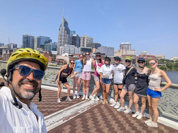 Smiling group of cyclists posing on a Nashville river bridge with the downtown skyline and a distinctive twin‑spired skyscraper behind them on a sunny day.