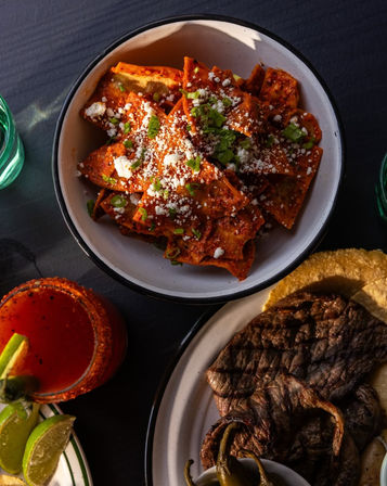 Bowl of spicy chilaquiles rojos sprinkled with queso fresco and scallions, paired with grilled steak on a tortilla, charred peppers, lime wedges and a chili-rimmed red cocktail.