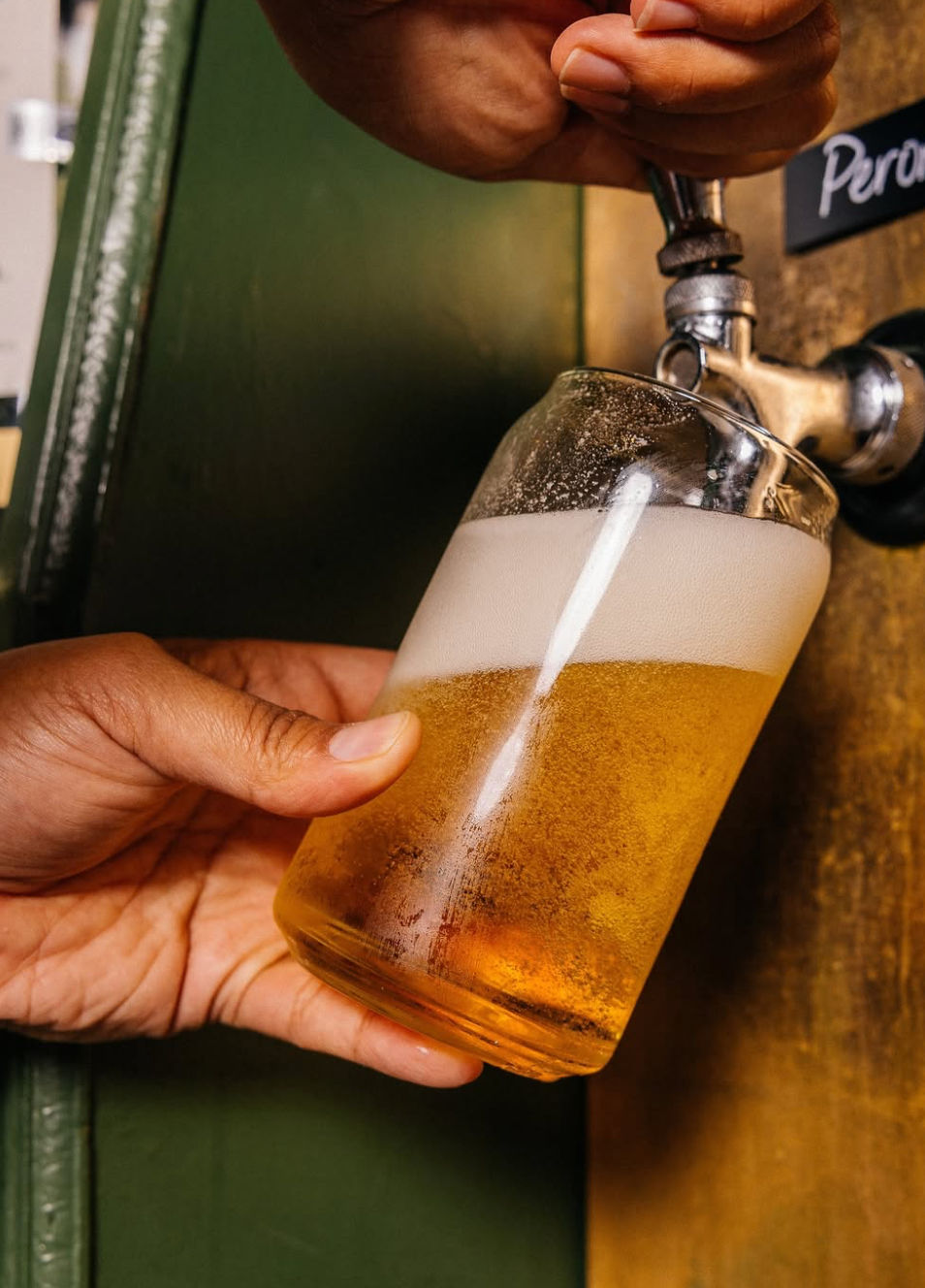 Close-up of a hand tilting a glass under a beer tap as cold golden draft beer fills it with a frothy white head, classic pub pour.