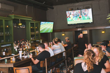 Lively neighborhood sports bar interior with patrons at a wooden bar and booths, bottles on green shelves, warm pendant lights, and two wall-mounted TVs showing a soccer match.