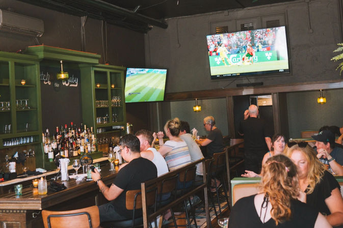 Lively neighborhood sports bar interior with patrons at a wooden bar and booths, bottles on green shelves, warm pendant lights, and two wall-mounted TVs showing a soccer match.