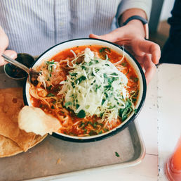 Hand holding an enamel bowl of Mexican-style red pozole topped with shredded cabbage and cilantro, served with crisp tostadas on a metal tray.