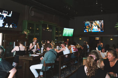 Lively dimly lit neighborhood sports bar interior with patrons seated at a wooden bar, bartenders serving drinks, and large TVs showing basketball and soccer