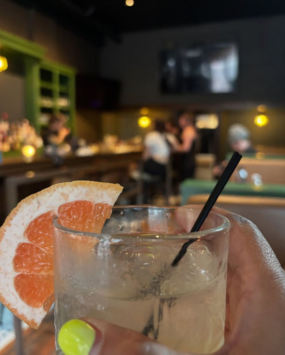 Close-up of a grapefruit-garnished cocktail in a rocks glass with a black straw and neon yellow manicure, blurred cozy bar interior with warm pendant lights and patrons in the background.