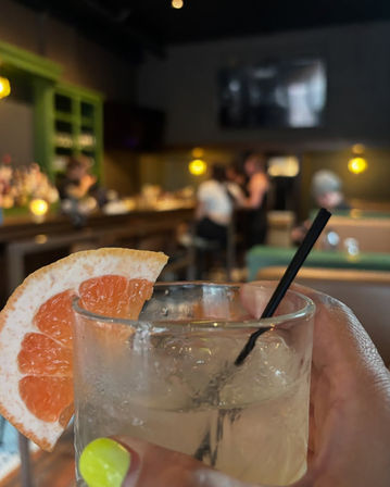 Close-up of a grapefruit-garnished cocktail in a rocks glass with a black straw and neon yellow manicure, blurred cozy bar interior with warm pendant lights and patrons in the background.