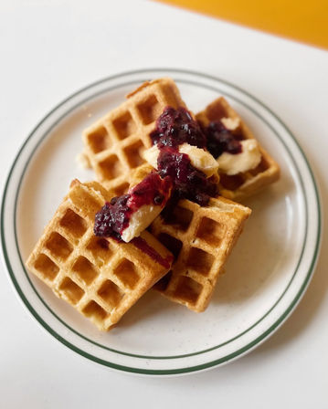 Crispy golden Belgian-style waffles stacked on a white plate with a green rim, topped with melting butter and glossy blueberry compote — brunch breakfast close-up
