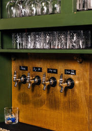 Four chrome beer taps on a warm brass panel with handwritten labels, patterned glasses on green shelves above and an empty rocks glass on the counter — craft beer taproom bar scene.