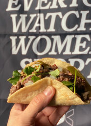 Hand holding a street-style corn tortilla taco filled with seasoned grilled beef, chopped onion, pickled jalapeño slices and fresh cilantro against a blurred black-and-white sign backdrop.