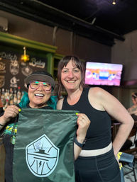 Two smiling friends in a casual bar holding a green drawstring bag with a white shield logo; one wears a cap, pink glasses and teal hair, the other wears a black tank top — lively pub interior with bottles and a TV in the background.