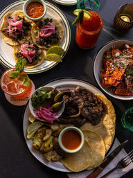 Top-down of a vibrant Mexican street-style spread: mushroom tacos with pickled red onions and salsa, a grilled steak plate with corn tortillas, roasted potatoes, pickled jalapeños and cilantro, a bowl of chilaquiles with cotija cheese, and two colorful cocktails garnished with lime and mint.