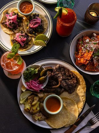 Top-down of a vibrant Mexican street-style spread: mushroom tacos with pickled red onions and salsa, a grilled steak plate with corn tortillas, roasted potatoes, pickled jalapeños and cilantro, a bowl of chilaquiles with cotija cheese, and two colorful cocktails garnished with lime and mint.