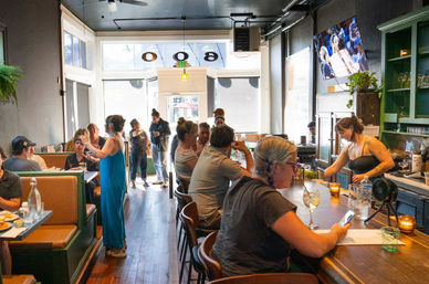 Bustling sunlit neighborhood cafe interior with wooden floors and green accents, patrons seated at booths and the bar while a bartender pours drinks near large front windows.