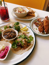 Plate of Mexican-style tacos topped with cilantro and diced onion, lime wedges and a bowl of chunky salsa, served with pickled onions, crispy breaded fritters, a bowl of chips with creamy corn, and a red iced drink.
