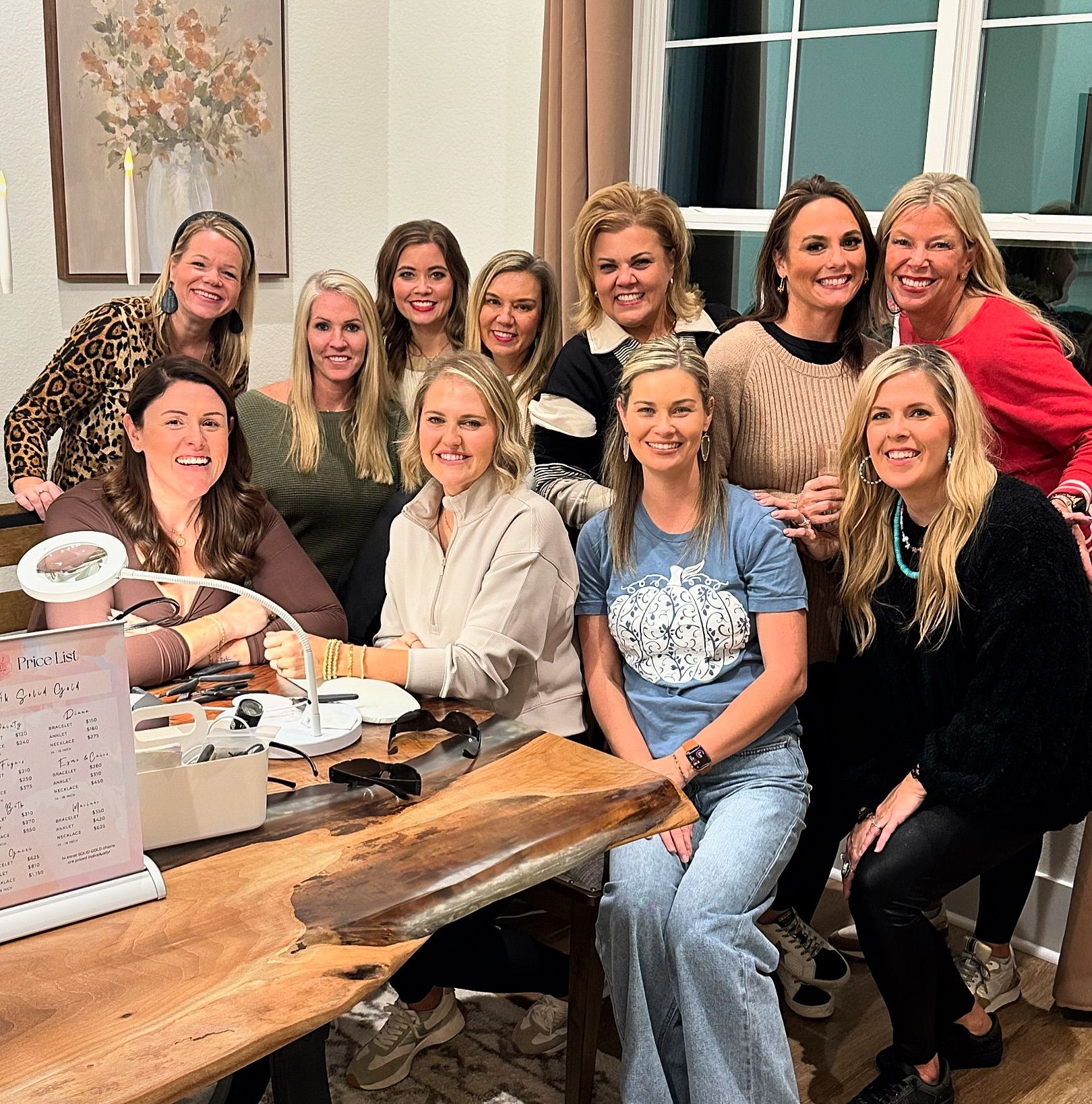 Smiling group of women gathered indoors around a live-edge wooden table with manicure tools and a price list, enjoying a cozy nail-party in a home setting.
