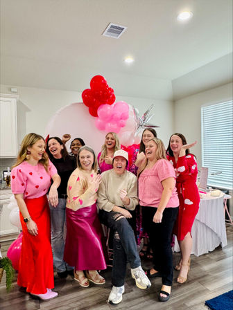 Group of smiling women posing at a bright home party by a pink and red heart-shaped balloon arch and silver star balloon over a circular backdrop, wearing Valentine-themed pink and red outfits in a kitchen/dining space.