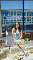 Smiling vendor at a sunny urban pop-up booth showcasing delicate gold bracelets, necklaces and rings on white displays with a small floral vase and price list, modern apartment building visible through large windows