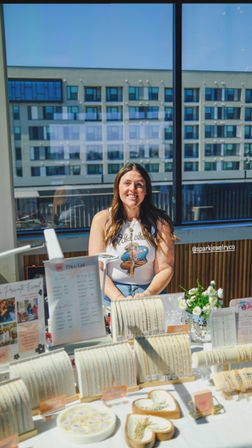 Smiling vendor at a sunny urban pop-up booth showcasing delicate gold bracelets, necklaces and rings on white displays with a small floral vase and price list, modern apartment building visible through large windows