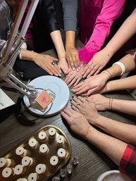 Manicure party: multiple manicured hands with rings and bracelets gathered around a nail lamp and trays of bracelet spools on a wooden table.