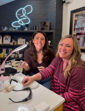 Two smiling women at a cozy nail salon manicure station, one receiving a gel manicure under a desk lamp with nail tools, a neon wall sign and shelves of beauty products in the background.