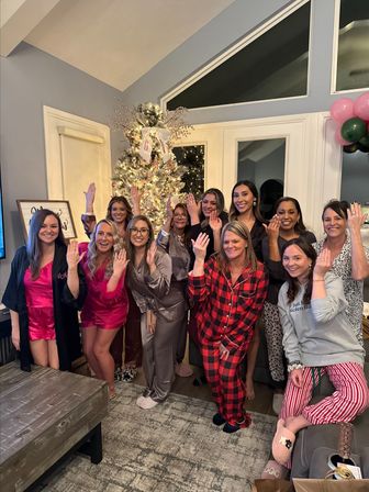 Group of women in festive pajamas and robes posing in a cozy living room holiday gathering, smiling and raising hands in front of a lit Christmas tree and balloons