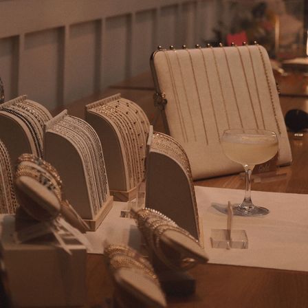 Warm-lit boutique table display of cream stands holding gold and silver bracelets and chains, a striped cream clutch with gold trim, and a coupe glass cocktail — stylish accessories scene.
