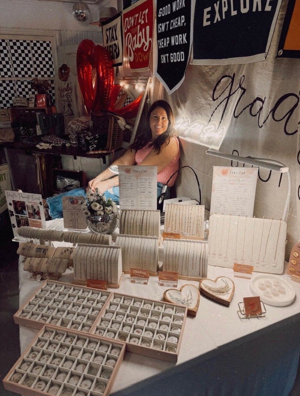 Smiling vendor at an indoor craft market booth showcasing handmade jewelry—organized trays of rings, bracelets and delicate necklaces on stands, with red heart balloons and inspirational banners in the cozy display.