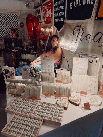 Smiling vendor at an indoor craft market booth showcasing handmade jewelry—organized trays of rings, bracelets and delicate necklaces on stands, with red heart balloons and inspirational banners in the cozy display.