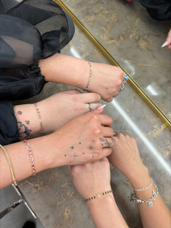 Close-up of four hands stacked on a marble countertop, showcasing colorful friendship bracelets, charm and chain bracelets, rings, and small star and floral hand tattoos — fashionable jewelry detail shot.