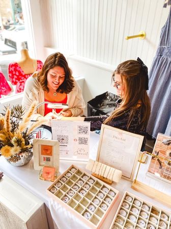 Two women at a bright, sunlit boutique pop-up table browsing organized jewelry display boxes, bracelet stand, dried florals, price list and QR payment signs.