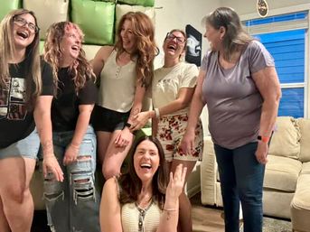Six women laughing together at a casual home gathering in a bright living room with a beige sofa and blue window — candid group of friends enjoying a fun celebration