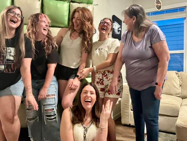 Six women laughing together at a casual home gathering in a bright living room with a beige sofa and blue window — candid group of friends enjoying a fun celebration