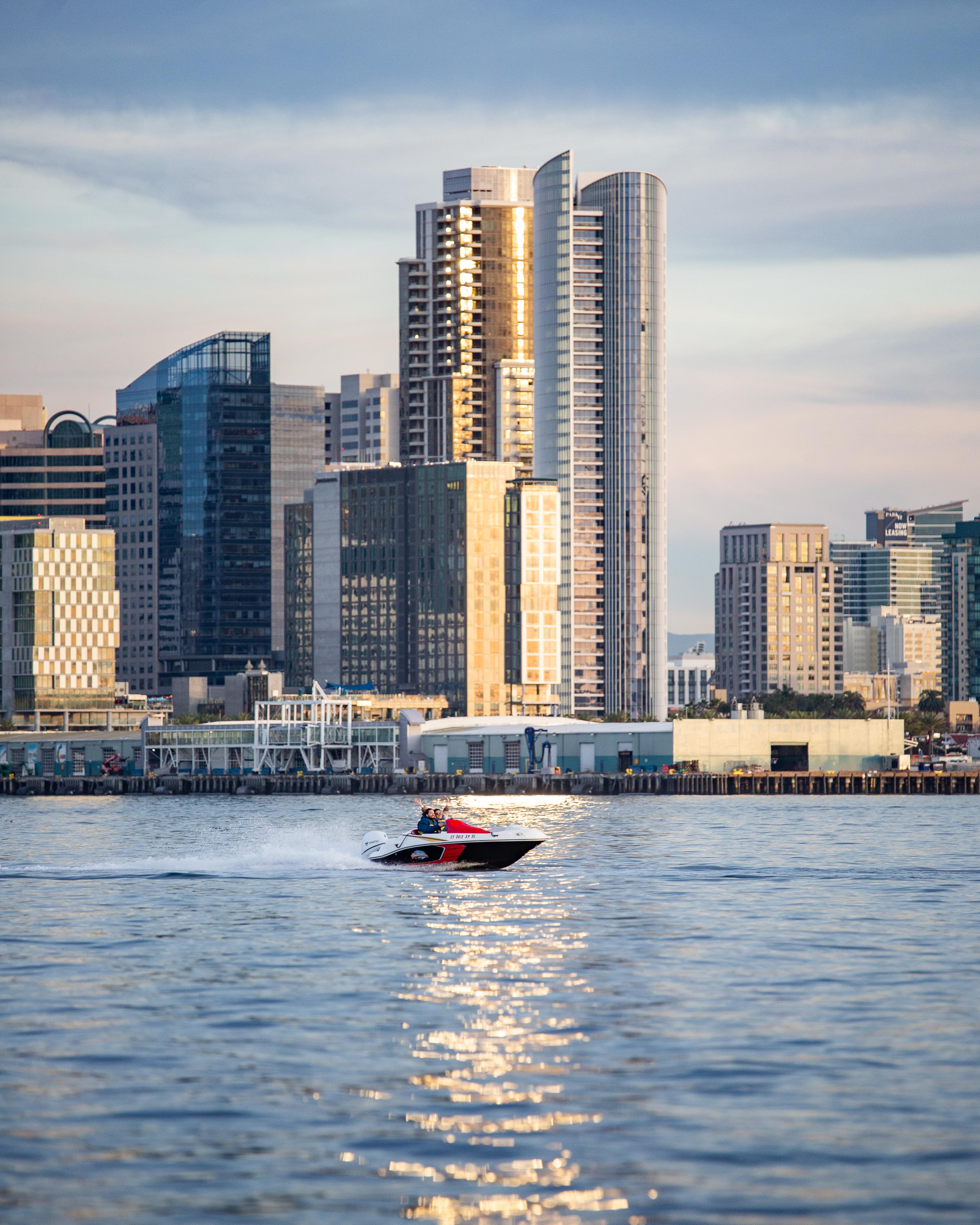 Red-and-white speedboat zips across a shimmering bay in front of a downtown waterfront skyline of glass high-rise towers glowing in golden sunset light.