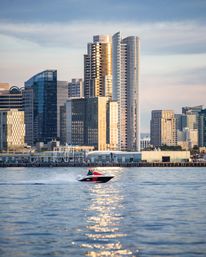 Red-and-white speedboat zips across a shimmering bay in front of a downtown waterfront skyline of glass high-rise towers glowing in golden sunset light.