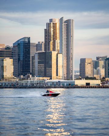 Red-and-white speedboat zips across a shimmering bay in front of a downtown waterfront skyline of glass high-rise towers glowing in golden sunset light.