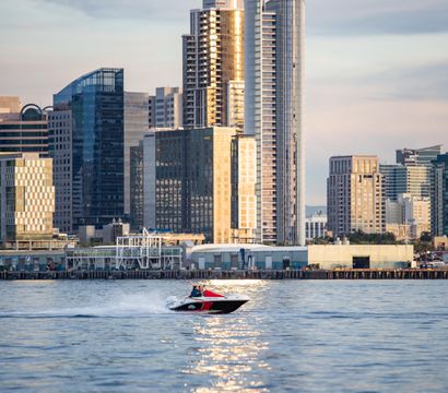 Red-and-white speedboat zips across a shimmering bay in front of a downtown waterfront skyline of glass high-rise towers glowing in golden sunset light.