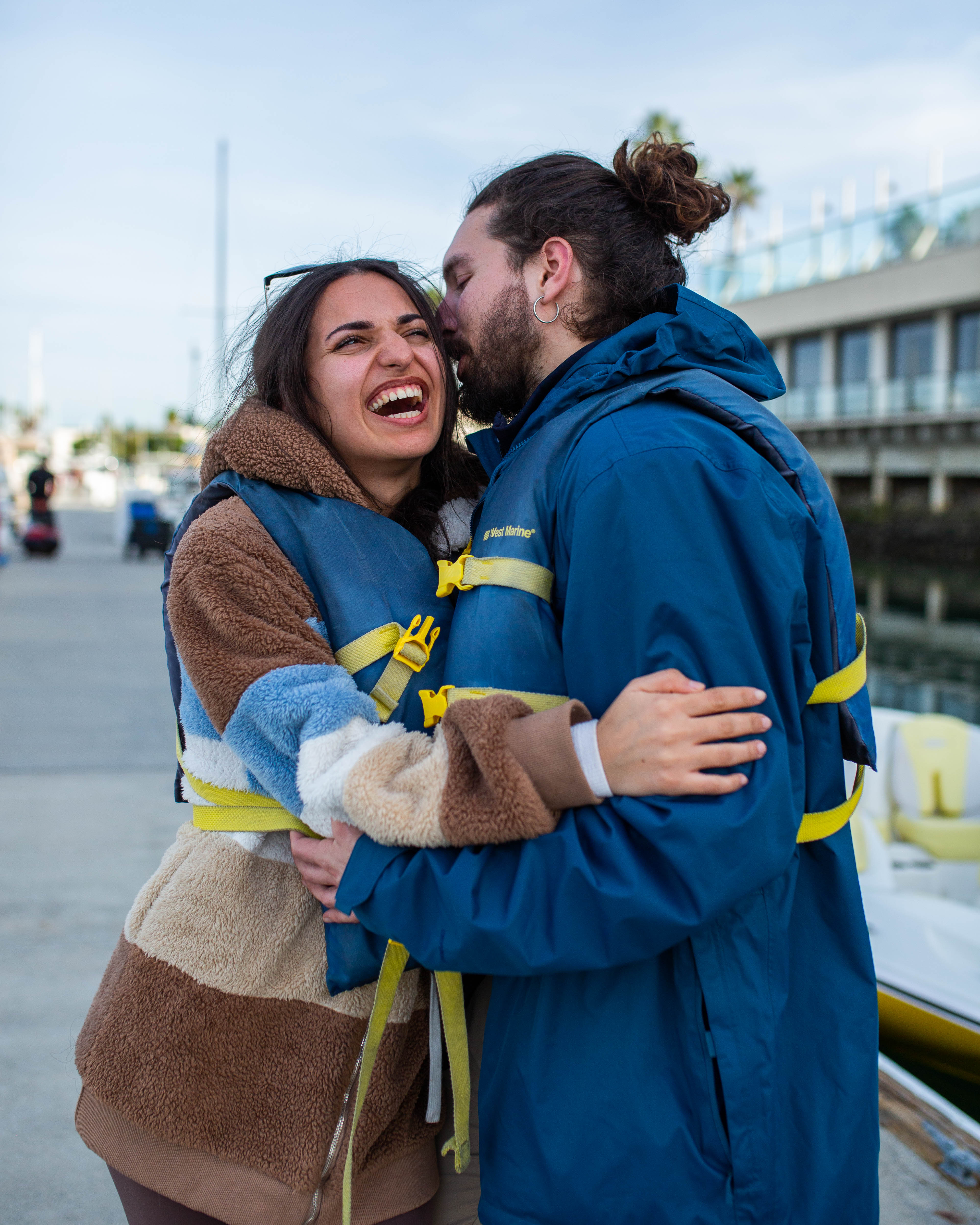 Playful couple wearing life jackets hugging and laughing on a marina dock beside moored boats and waterfront buildings