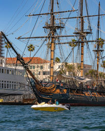 Historic tall ship with wooden masts and intricate rigging docked at a sunny palm-lined harbor, a small yellow motorboat with two passengers cruising in the blue water foreground.
