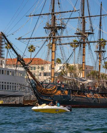 Historic tall ship with wooden masts and intricate rigging docked at a sunny palm-lined harbor, a small yellow motorboat with two passengers cruising in the blue water foreground.