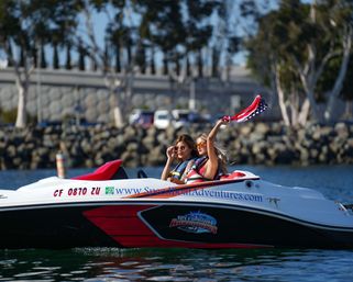 Two women in sunglasses and life jackets riding a red-and-white speedboat near a rocky marina, one waving an American-flag bandana over the water on a sunny coastal harbor day.
