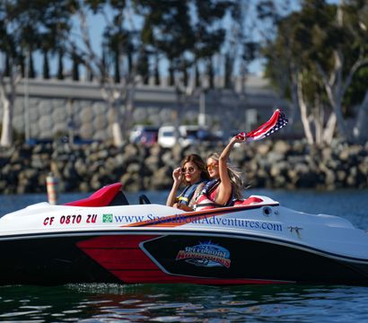 Two women in sunglasses and life jackets riding a red-and-white speedboat near a rocky marina, one waving an American-flag bandana over the water on a sunny coastal harbor day.