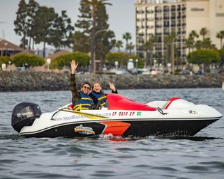 Two people in life jackets waving from a red-and-white speedboat cruising in a palm-lined harbor near waterfront hotels