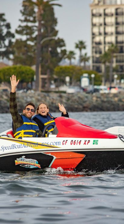 Two people in life jackets waving from a red-and-white speedboat cruising in a palm-lined harbor near waterfront hotels